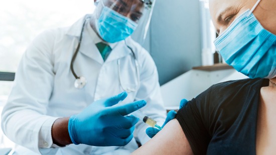 A physician wearing a face shield and surgical mask administers a vaccine to a patient in a surgical mask.