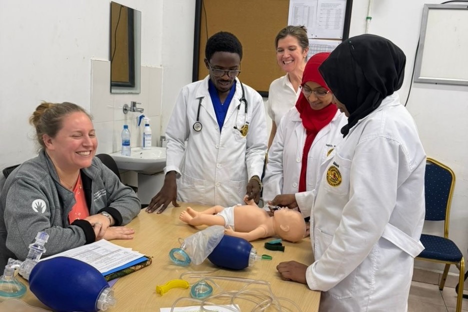 A medical team stands around a simulation doll on a table